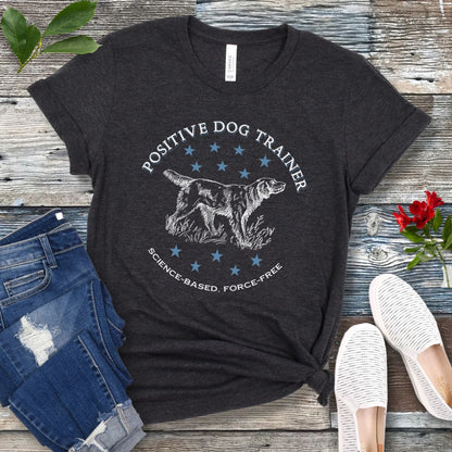 A dark grey t-shirt with a vintage dog illustration and text that reads 'POSITIVE DOG TRAINER SCIENCE-BASED FORCE-FREE', displayed on a wooden background with jeans and white shoes.