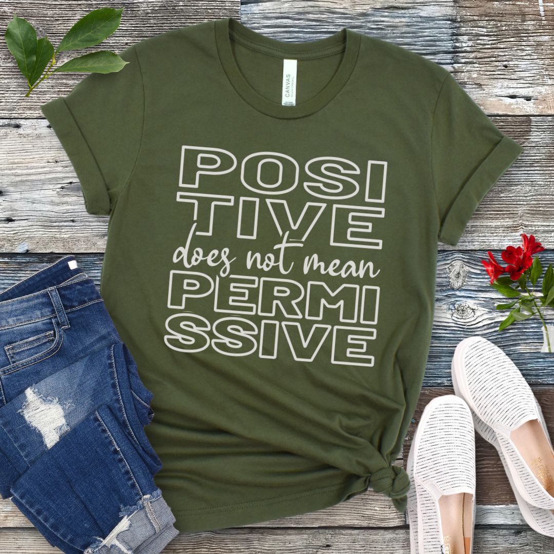 Military green dog training t-shirt with 'POSITIVE does not mean PERMISSIVE' typography text on a wooden surface with jeans and shoes.