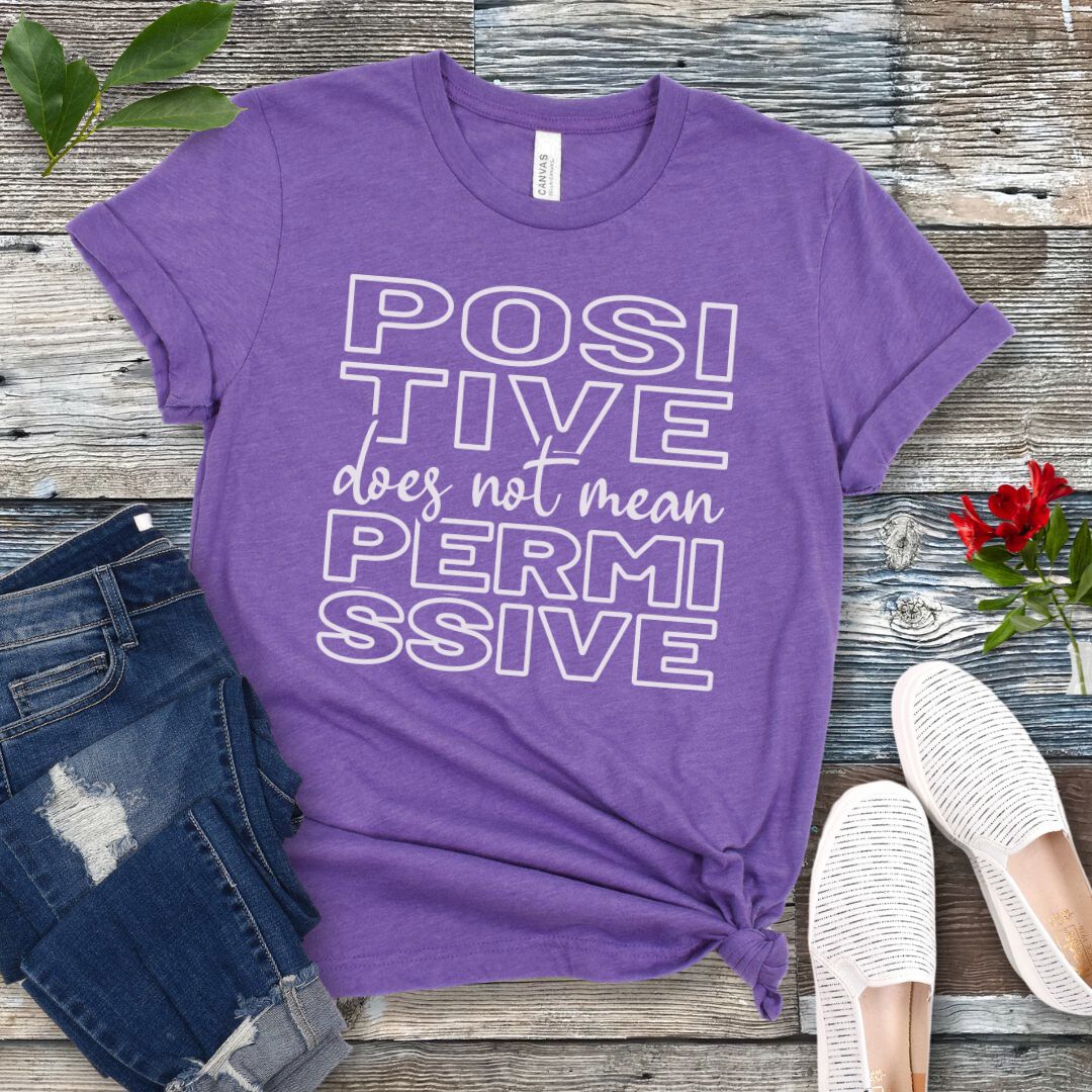 Purple dog training t-shirt with 'POSITIVE does not mean PERMISSIVE' typography text on a wooden surface with jeans and shoes.