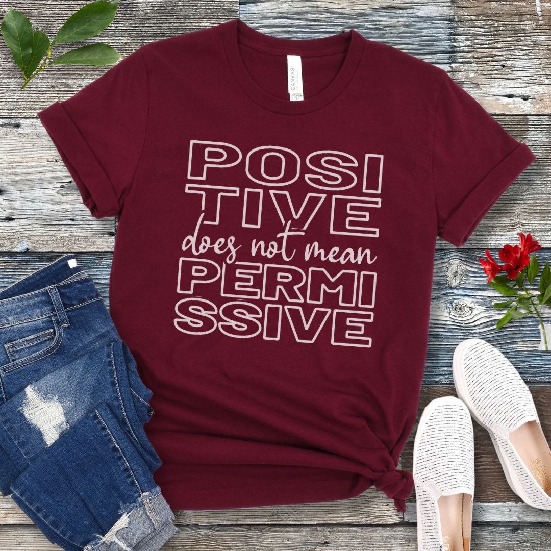 Maroon dog training t-shirt with 'POSITIVE does not mean PERMISSIVE' typography text on a wooden surface with jeans and shoes.