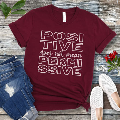 Maroon dog training t-shirt with 'POSITIVE does not mean PERMISSIVE' typography text on a wooden surface with jeans and shoes.