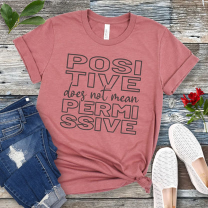 Mauve dog training t-shirt with 'POSITIVE does not mean PERMISSIVE' typography text on a wooden surface with jeans and shoes.