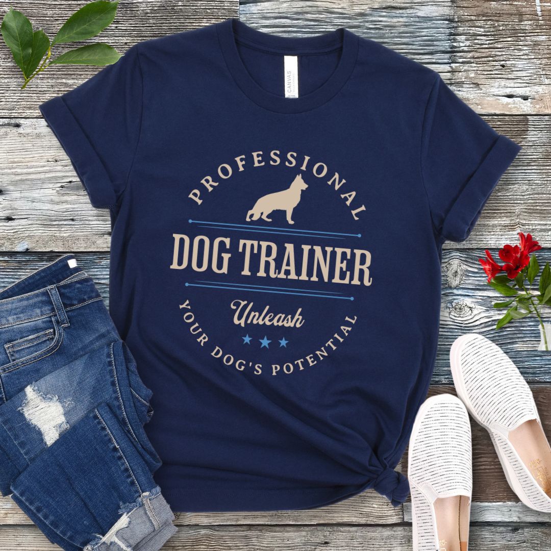 A navy blue unisex t-shirt with the text 'PROFESSIONAL DOG TRAINER, Unleash Your Dog's Potential' and a custom breed dog silhouette, in vintage badge style, laid out flat with jeans and white shoes on a wooden surface.