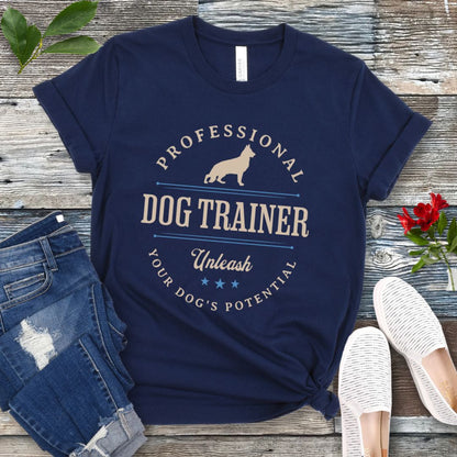 A navy blue unisex t-shirt with the text 'PROFESSIONAL DOG TRAINER, Unleash Your Dog's Potential' and a custom breed dog silhouette, in vintage badge style, laid out flat with jeans and white shoes on a wooden surface.