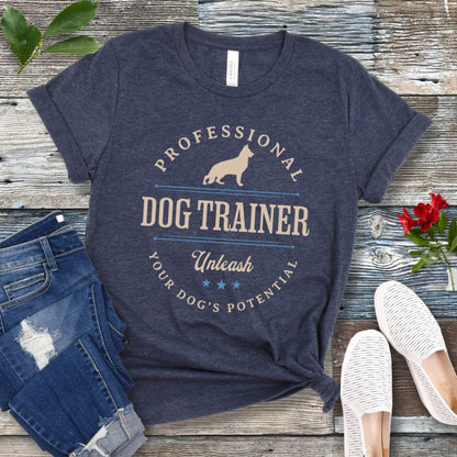 A heather navy unisex t-shirt with the text 'PROFESSIONAL DOG TRAINER, Unleash Your Dog's Potential' and a custom breed dog silhouette, in vintage badge style, laid out flat with jeans and white shoes on a wooden surface.