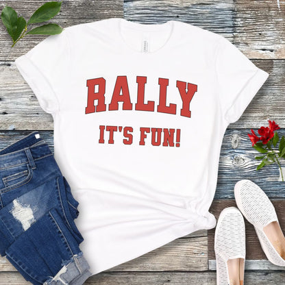 A white Rally dog obedience t-shirt with 'RALLY IT'S FUN!' printed in red college style lettering, displayed with blue jeans and white shoes on a wooden background.