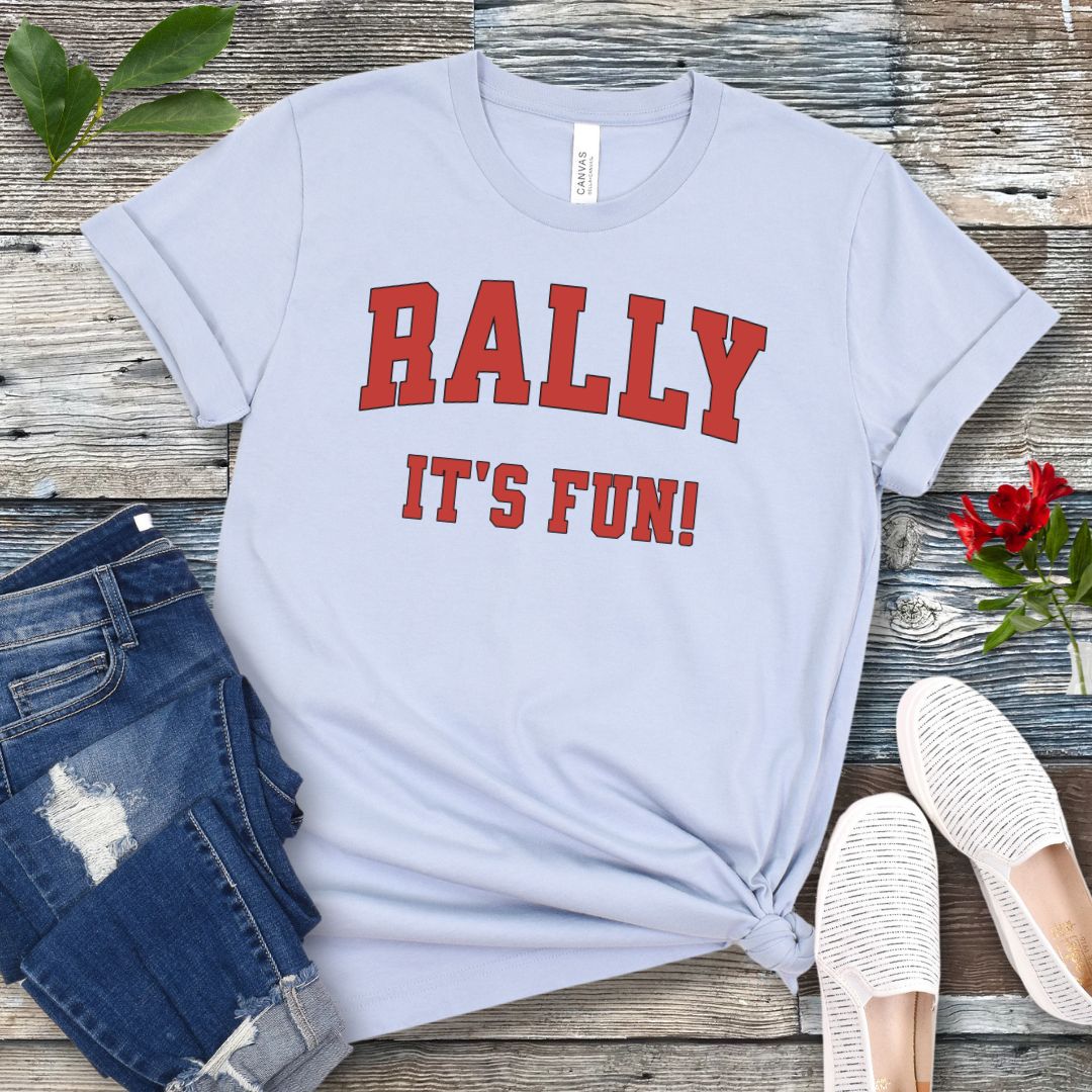 A light blue Rally dog obedience t-shirt with 'RALLY IT'S FUN!' printed in red college style lettering, displayed with blue jeans and white shoes on a wooden background.