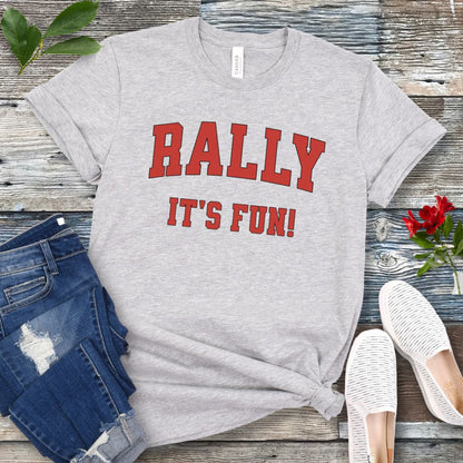 A grey Rally dog obedience t-shirt with 'RALLY IT'S FUN!' printed in red college style lettering, displayed with blue jeans and white shoes on a wooden background.