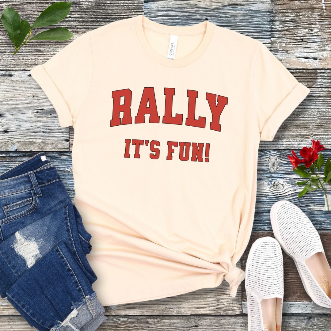A beige Rally dog obedience t-shirt with 'RALLY IT'S FUN!' printed in red college style lettering, displayed with blue jeans and white shoes on a wooden background.