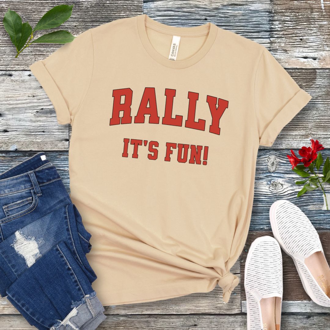 A cream colored Rally dog obedience t-shirt with 'RALLY IT'S FUN!' printed in red college style lettering, displayed with blue jeans and white shoes on a wooden background.