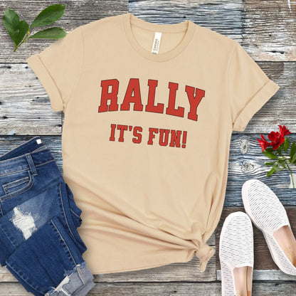 A cream colored Rally dog obedience t-shirt with 'RALLY IT'S FUN!' printed in red college style lettering, displayed with blue jeans and white shoes on a wooden background.