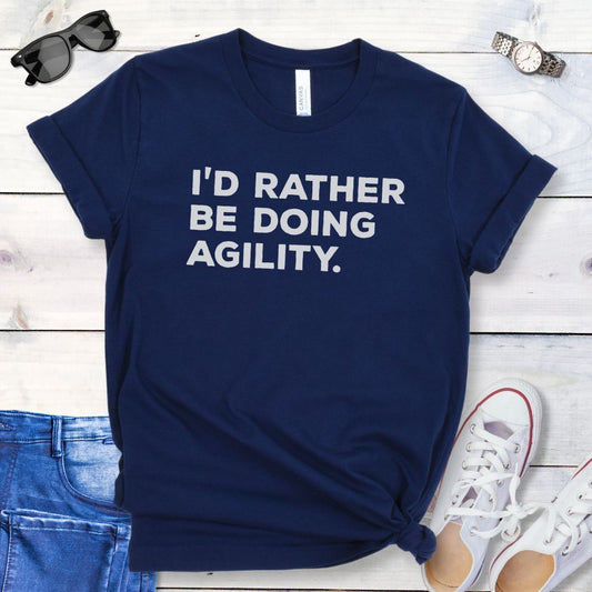 Navy blue dog sports t-shirt with bold 'I'd Rather Be Doing Agility' text on a wooden surface with jeans and shoes.