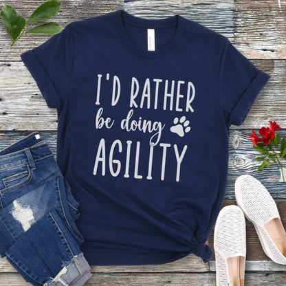 Navy blue dog sports t-shirt with 'I'd Rather be doing Agility' text on a wooden surface with jeans and shoes.