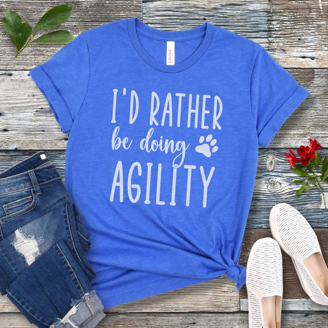 Royal blue dog sports t-shirt with 'I'd Rather be doing Agility' text on a wooden surface with jeans and shoes.