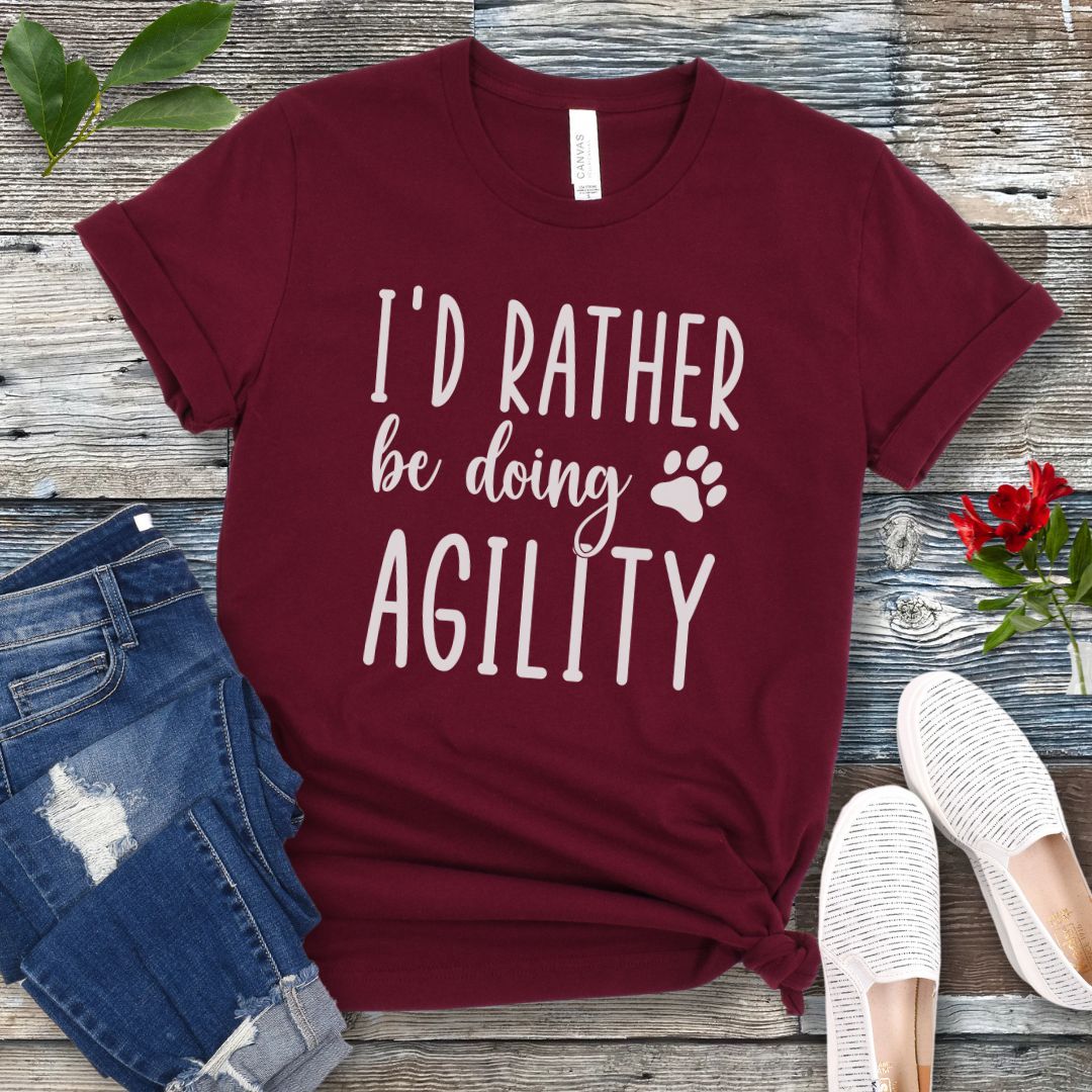 Maroon dog sports t-shirt with 'I'd Rather be doing Agility' text on a wooden surface with jeans and shoes.