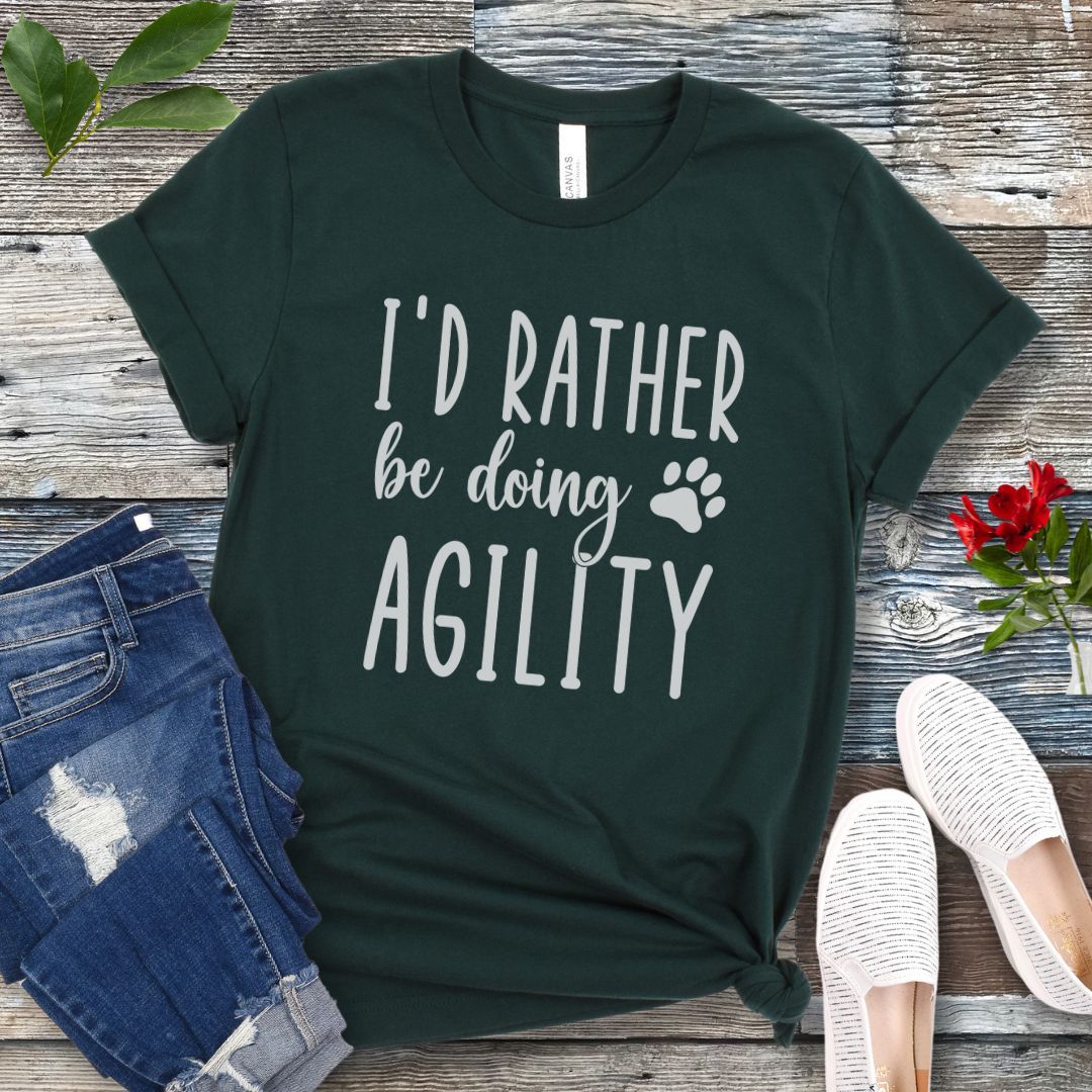 Dark green dog sports t-shirt with 'I'd Rather be doing Agility' text on a wooden surface with jeans and shoes.
