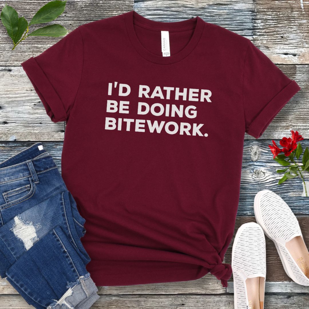 Maroon t-shirt with 'I'd Rather Be Doing Bitework' text on a wooden surface with jeans and shoes.