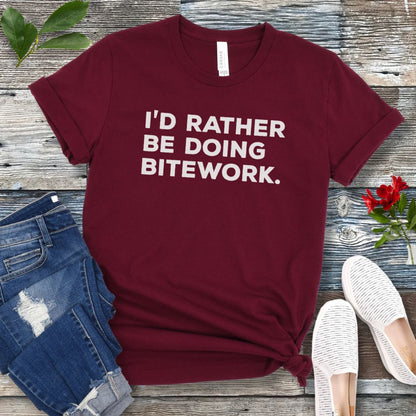 Maroon t-shirt with 'I'd Rather Be Doing Bitework' text on a wooden surface with jeans and shoes.