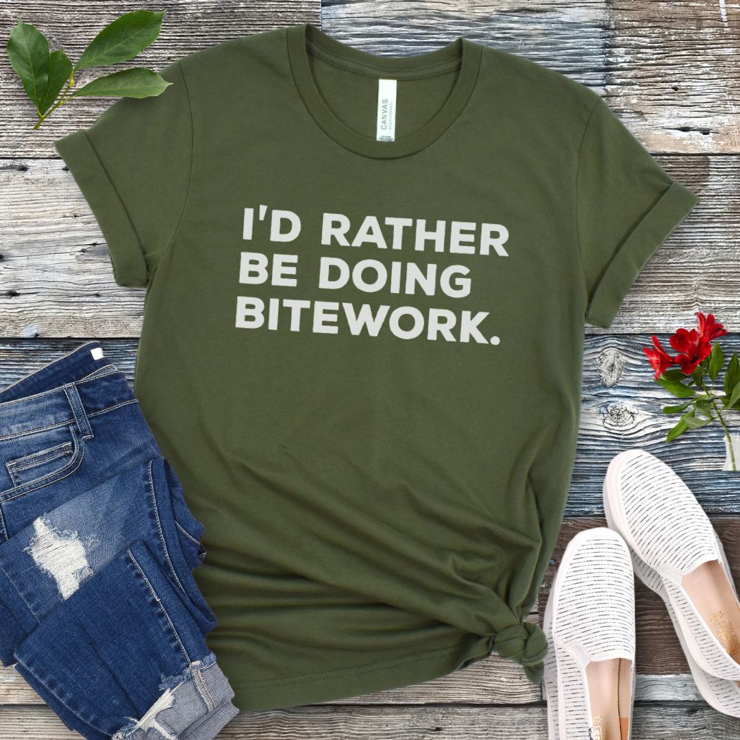 Military green t-shirt with 'I'd Rather Be Doing Bitework' text on a wooden surface with jeans and shoes.