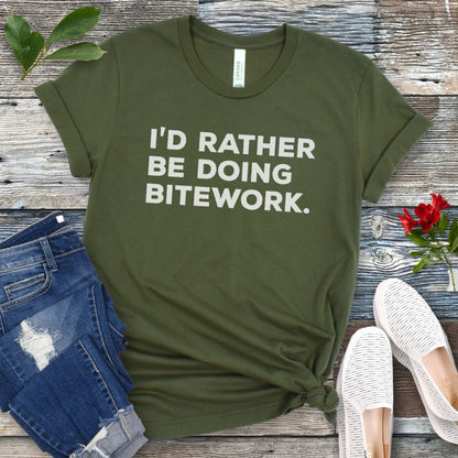 Military green t-shirt with 'I'd Rather Be Doing Bitework' text on a wooden surface with jeans and shoes.