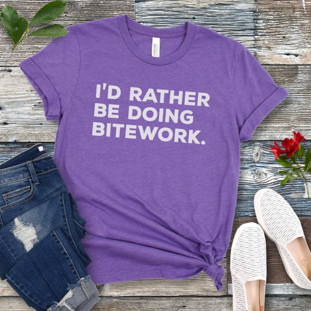 Purple t-shirt with 'I'd rather be doing bitework' text on a wooden surface with jeans and shoes.