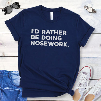 Navy t-shirt for dog scent trainers with the humorous text 'I'd Rather Be Doing Nosework' in bold, blocky lettering, shown  on a wooden surface with casual items.