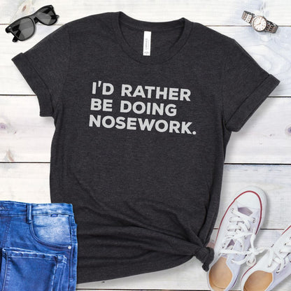 Dark grey t-shirt for dog scent trainers with the humorous text 'I'd Rather Be Doing Nosework' in bold, blocky lettering, shown  on a wooden surface with casual items.