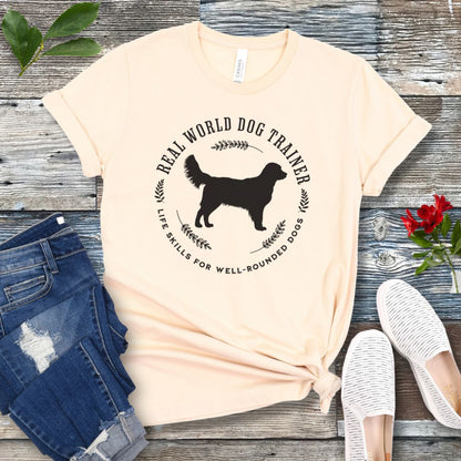 A beige dog training t-shirt with a graphic of a Golden Retriever dog and the text 'REAL WORLD DOG TRAINER Life skills for well-rounded dogs'. The t-shirt is presented flat with a pair of blue jeans and white sneakers, suggesting a casual setting.