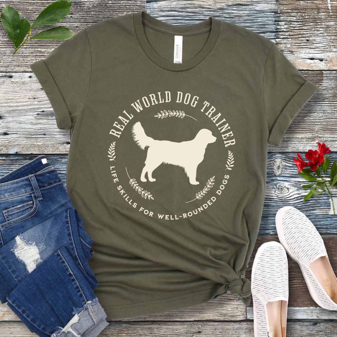 A military green dog training t-shirt with a graphic of a Golden Retriever dog and the text 'REAL WORLD DOG TRAINER Life skills for well-rounded dogs'. The t-shirt is presented flat with a pair of blue jeans and white sneakers, suggesting a casual setting.