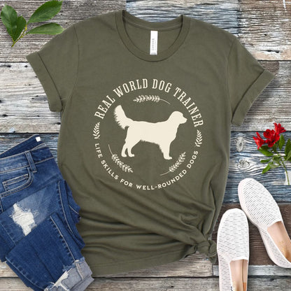 A military green dog training t-shirt with a graphic of a Golden Retriever dog and the text 'REAL WORLD DOG TRAINER Life skills for well-rounded dogs'. The t-shirt is presented flat with a pair of blue jeans and white sneakers, suggesting a casual setting.