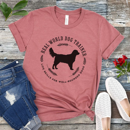 A mauve dog training t-shirt with a graphic of a Golden Retriever dog and the text 'REAL WORLD DOG TRAINER Life skills for well-rounded dogs'. The t-shirt is presented flat with a pair of blue jeans and white sneakers, suggesting a casual setting.