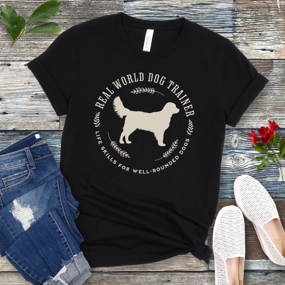 A black dog training t-shirt with a graphic of a Golden Retriever dog and the text 'REAL WORLD DOG TRAINER Life skills for well-rounded dogs'. The t-shirt is presented flat with a pair of blue jeans and white sneakers, suggesting a casual setting.
