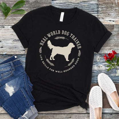 A black dog training t-shirt with a graphic of a Golden Retriever dog and the text 'REAL WORLD DOG TRAINER Life skills for well-rounded dogs'. The t-shirt is presented flat with a pair of blue jeans and white sneakers, suggesting a casual setting.
