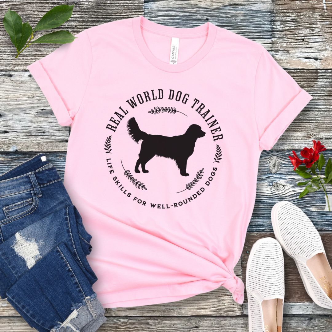 A pink dog training t-shirt with a graphic of a Golden Retriever dog and the text 'REAL WORLD DOG TRAINER Life skills for well-rounded dogs'. The t-shirt is presented flat with a pair of blue jeans and white sneakers, suggesting a casual setting.