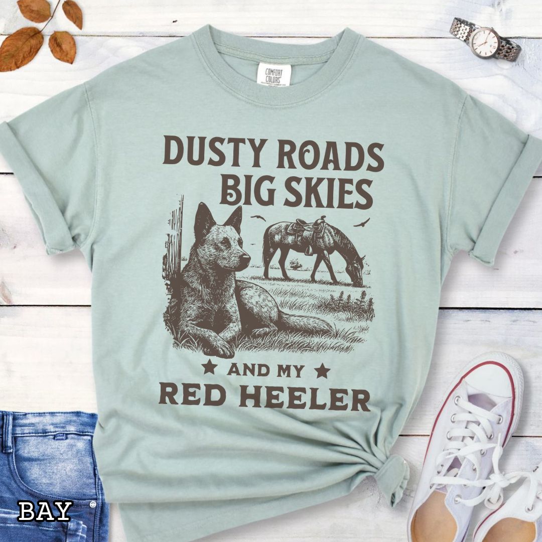 Light green Comfort Colors Red Heeler t-shirt featuring a Western-style design with grazing horse, rustic fence, and an Cattle Dog illustration; text reads “Dusty Roads, Big Skies, and My Red Heeler.”