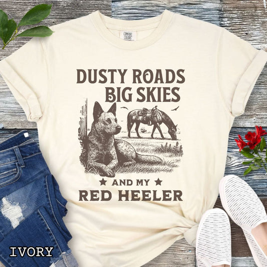 Ivory Comfort Colors Red Heeler t-shirt featuring a Western-style design with grazing horse, rustic fence, and an Cattle Dog illustration; text reads “Dusty Roads, Big Skies, and My Red Heeler.”