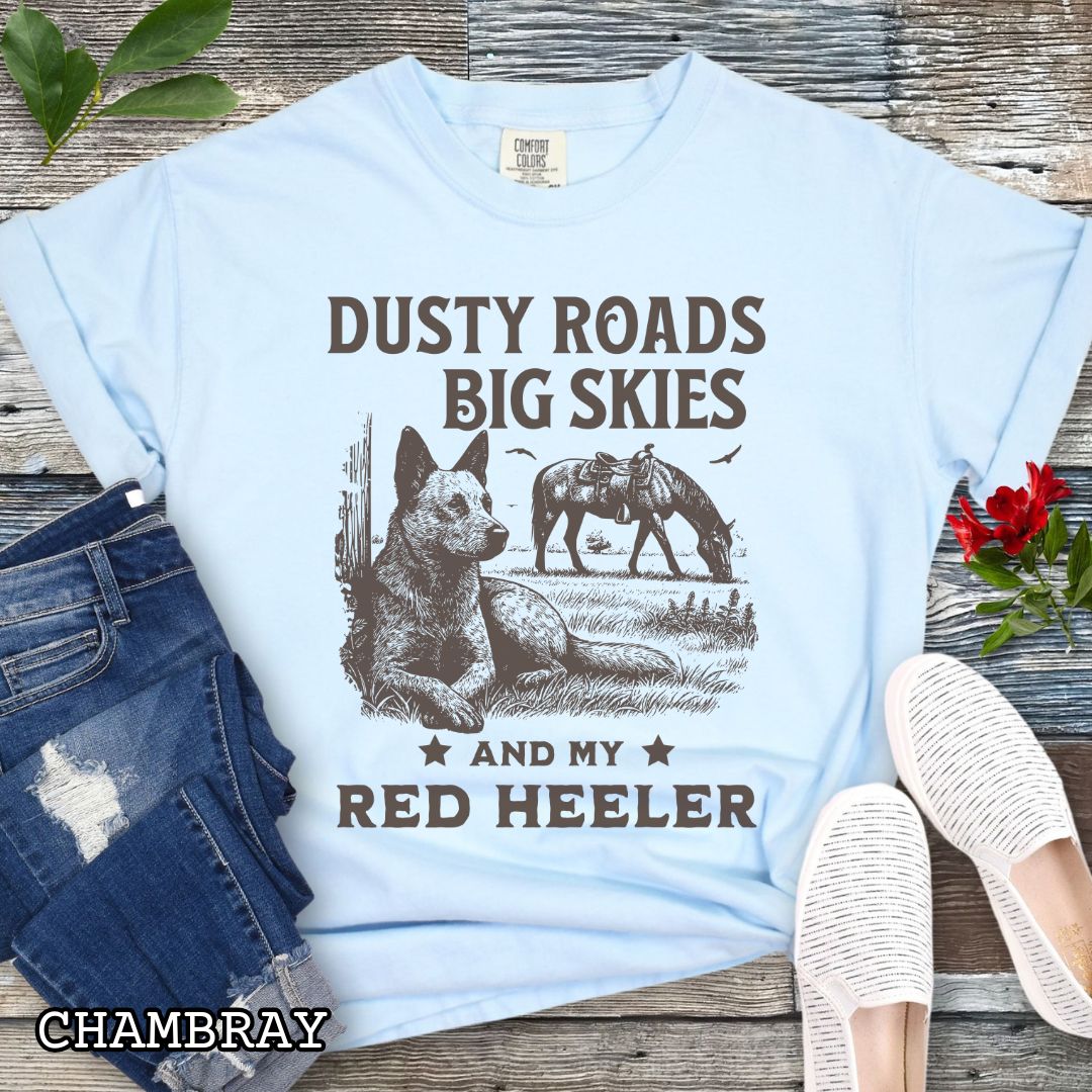 Light blue Comfort Colors Red Heeler t-shirt featuring a Western-style design with grazing horse, rustic fence, and a Cattle Dog illustration; text reads “Dusty Roads, Big Skies, and My Red Heeler.”