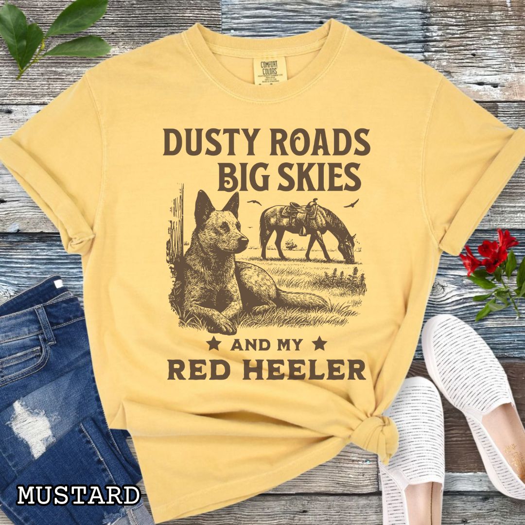 Mustard-yellow Comfort Colors Red Heeler t-shirt featuring a Western-style design with grazing horse, rustic fence, and a Cattle Dog illustration; text reads “Dusty Roads, Big Skies, and My Red Heeler.”