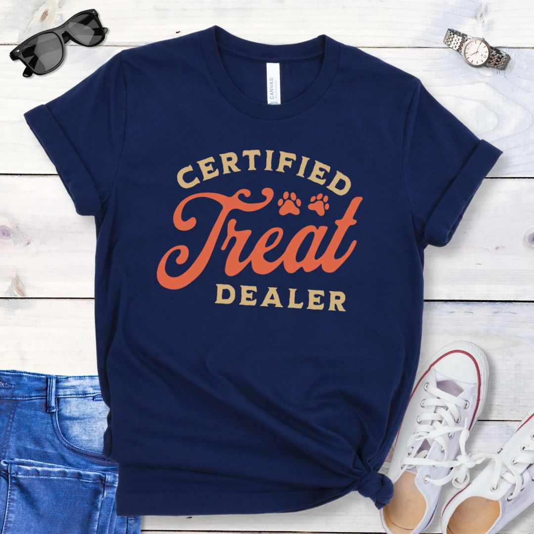 Navy dog lover t-shirt with the text 'CERTIFIED TREAT DEALER' printed in a retro vintage style font, accompanied by two paw prints. The shirt is presented on a wooden surface with a pair of blue jeans and white shoes in the background.
