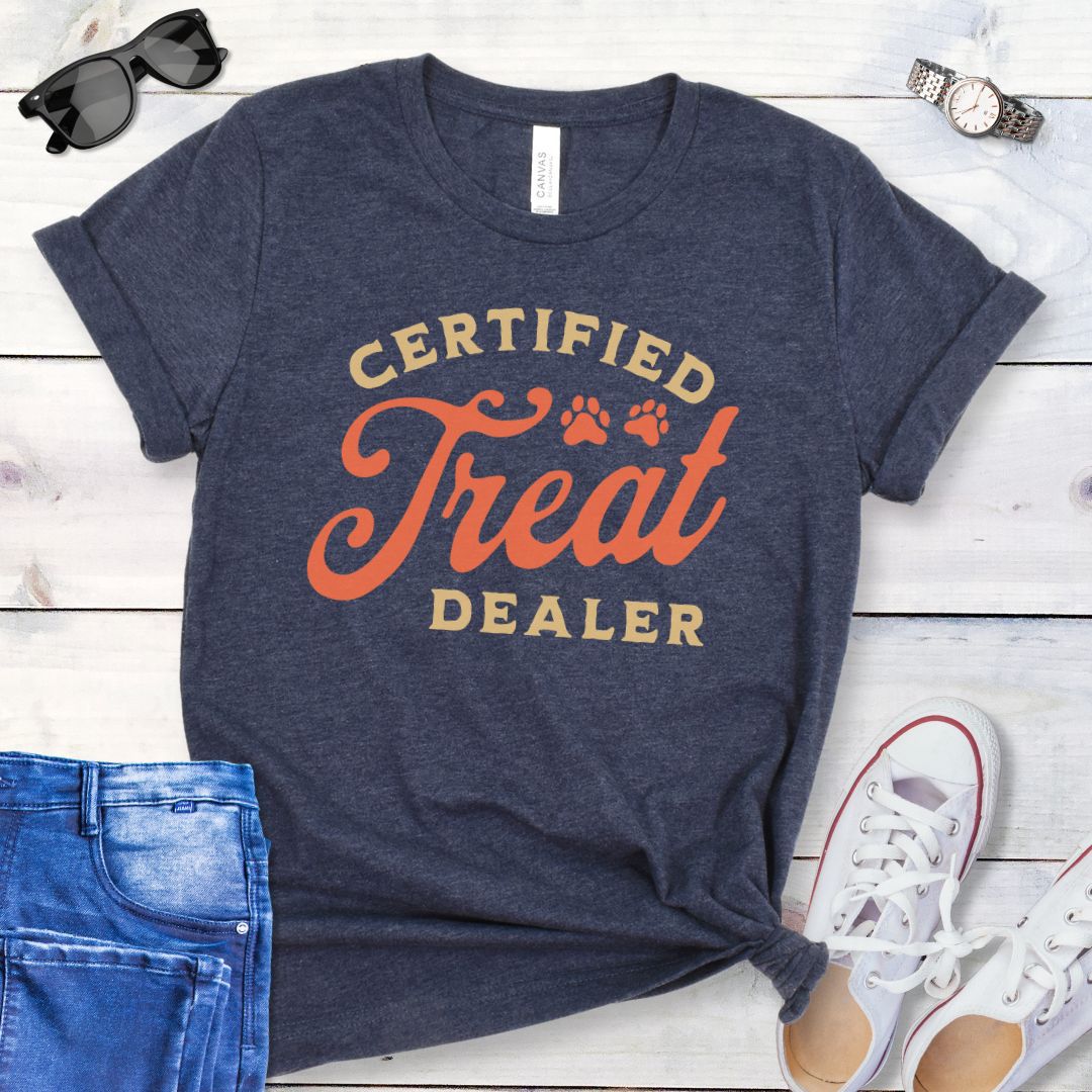 Heather navy dog lover t-shirt with the text 'CERTIFIED TREAT DEALER' printed in a retro vintage style font, accompanied by two paw prints. The shirt is presented on a wooden surface with a pair of blue jeans and white shoes in the background.