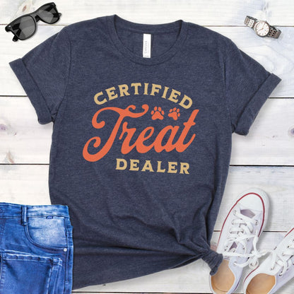 Heather navy dog lover t-shirt with the text 'CERTIFIED TREAT DEALER' printed in a retro vintage style font, accompanied by two paw prints. The shirt is presented on a wooden surface with a pair of blue jeans and white shoes in the background.