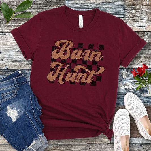 A maroon-colored t-shirt with the text 'Barn Hunt' in a vintage-style font over a checkerboard pattern, displayed on a wooden background with a pair of blue jeans and white shoes.