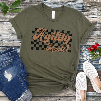 A military greenn dog agility t-shirt with 'Agility Mode' written in a retro stylized font over a checkerboard pattern, placed on a wooden surface, accompanied by jeans and white shoes.