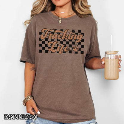 Woman wearing a brown t-shirt with checkerboard, paw, and 'Tracking Life' text in retro font.