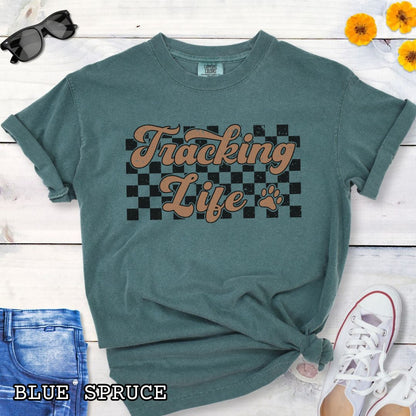 Green t-shirt with checkerboard, paw, and 'Tracking Life' text in retro font, presented on a wooden surface with sunglasses and jeans.