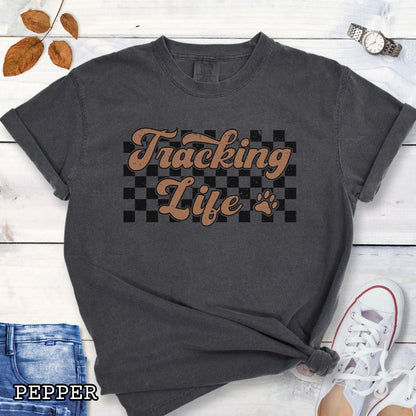 Dark grey t-shirt with checkerboard, paw, and 'Tracking Life' text in retro font, presented on a wooden surface with sunglasses and jeans.