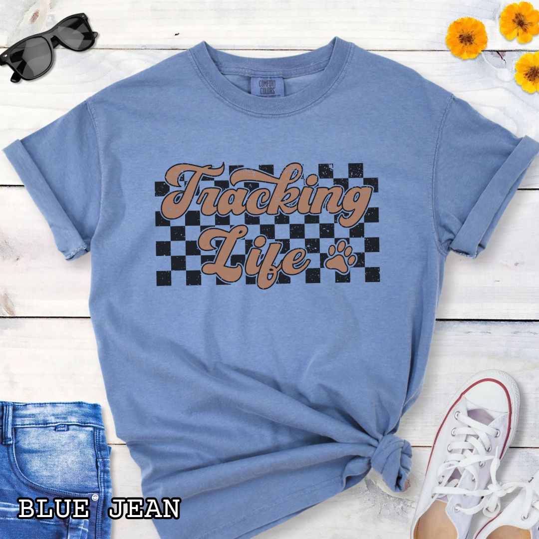 Blue t-shirt with checkerboard, paw, and 'Tracking Life' text in retro font, presented on a wooden surface with sunglasses and jeans.