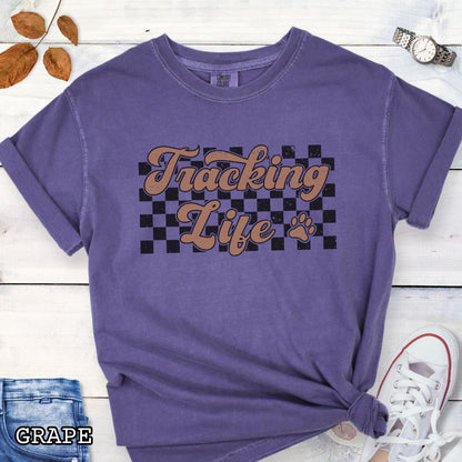Purple t-shirt with checkerboard, paw, and 'Tracking Life' text in retro font, presented on a wooden surface with sunglasses and jeans.