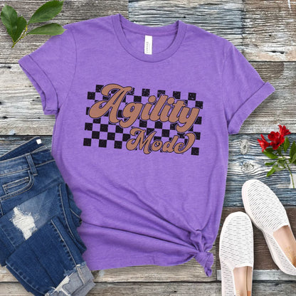 A purple dog agility t-shirt with 'Agility Mode' written in a retro stylized font over a checkerboard pattern, placed on a wooden surface, accompanied by jeans and white shoes.