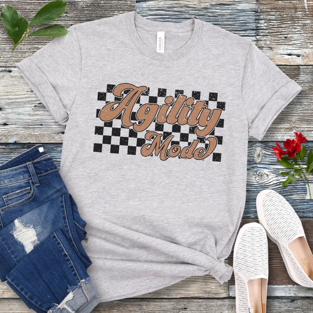 A grey dog agility t-shirt with 'Agility Mode' written in a retro stylized font over a checkerboard pattern, placed on a wooden surface, accompanied by jeans and white shoes.